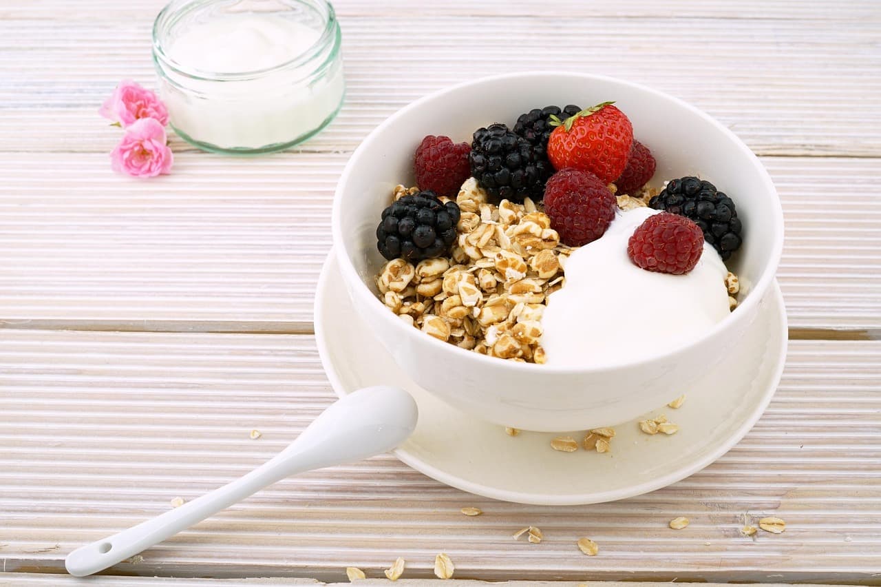 Bowl of oats, fresh berries, and yogurt on a white wood table, with a small jar of yogurt and a sprig of pink roses