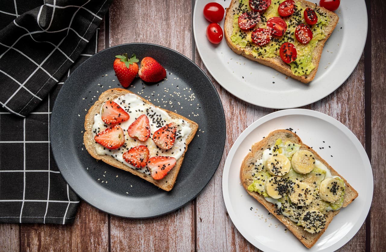 Three open-faced toasts on plates seen from above on a dark wooden table — strawberry and seed, tomato and avocado, banana and avocado — with a checked tea towel to the side