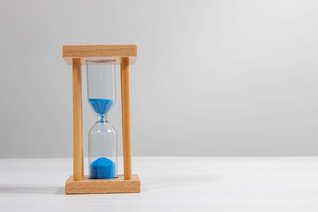 A wooden-frame hourglass with bright blue sand mid-pour on a clean white surface in soft daylight