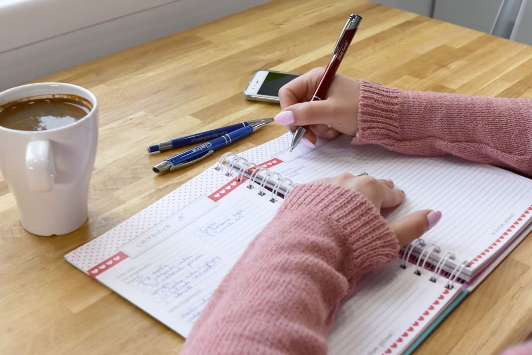 A woman in a cozy knitted sweater writing in a notebook at a wooden table beside a cup of coffee in warm morning light