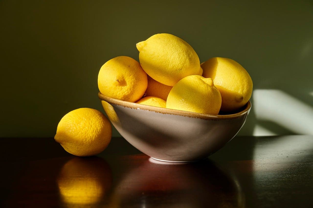 Fresh lemons in a shallow bowl on a pale surface in warm side light, soft shadows, still-life composition