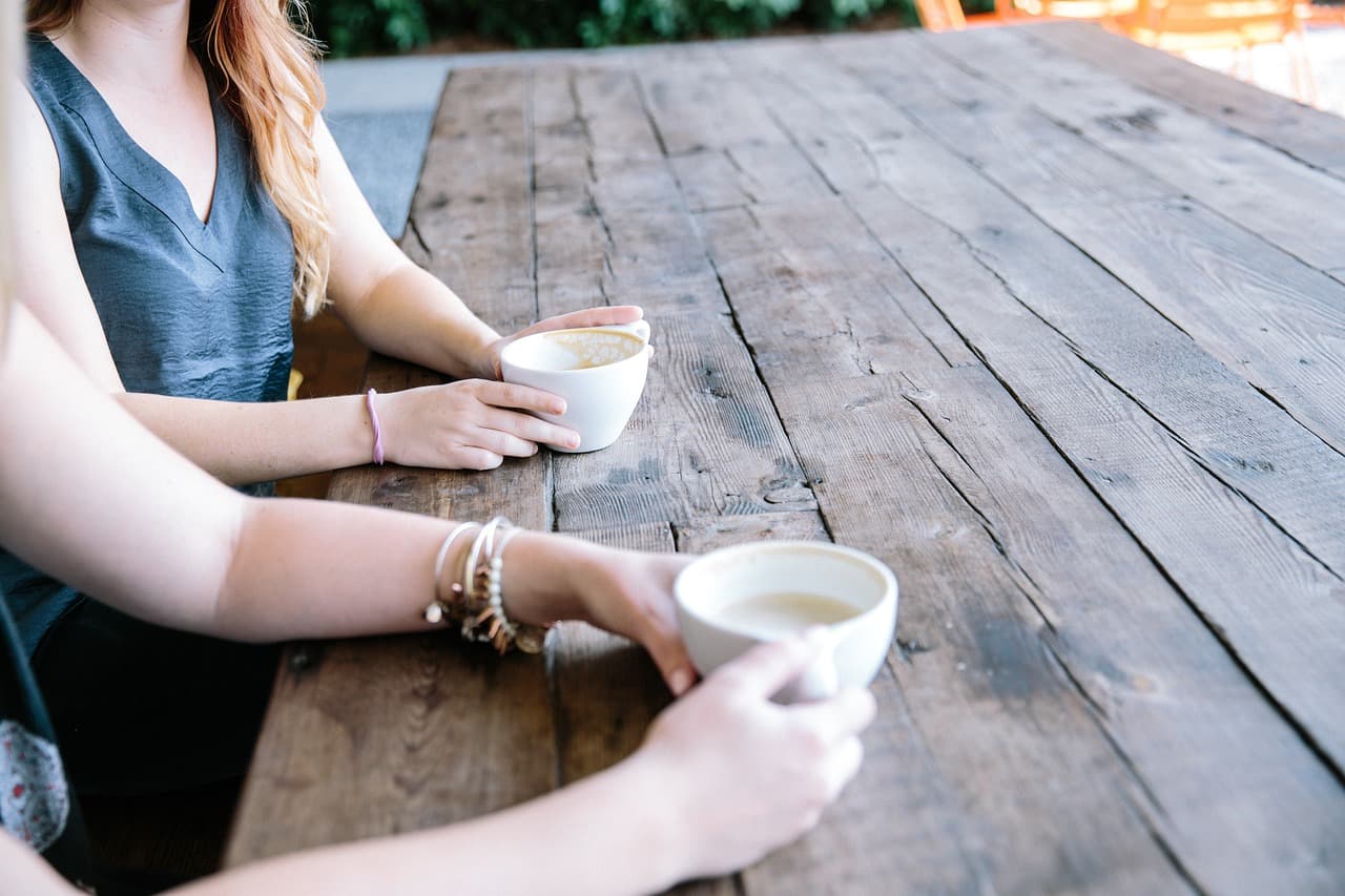 Two people sitting at a wooden table with coffee mugs, talking in relaxed natural daylight