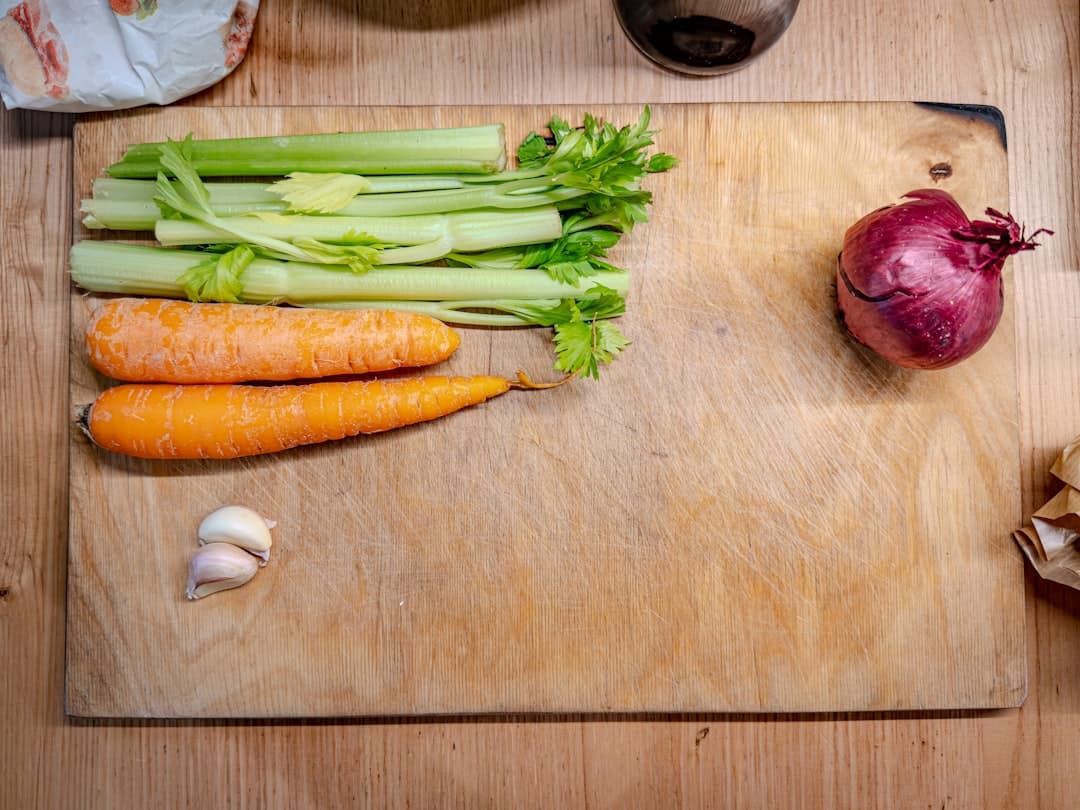 Celery, carrots, an onion, and garlic cloves waiting to be chopped on a well-used wooden board in a home kitchen