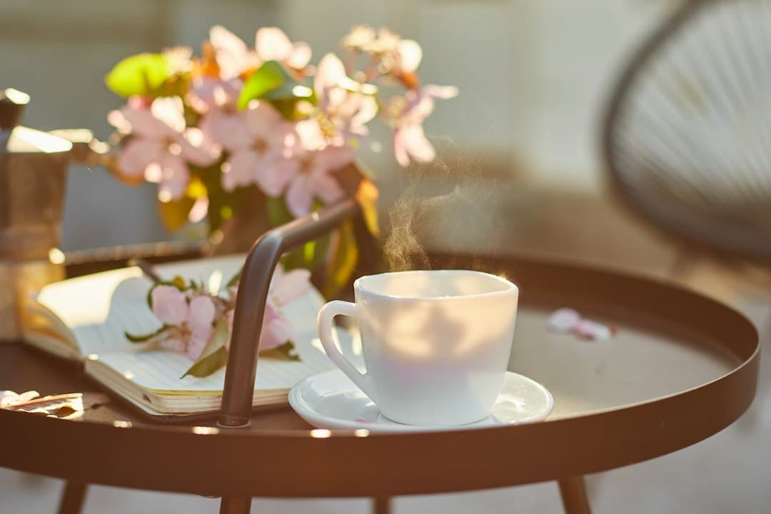 A steaming cup on a tray beside an open book and pink blossoms on a sunny terrace