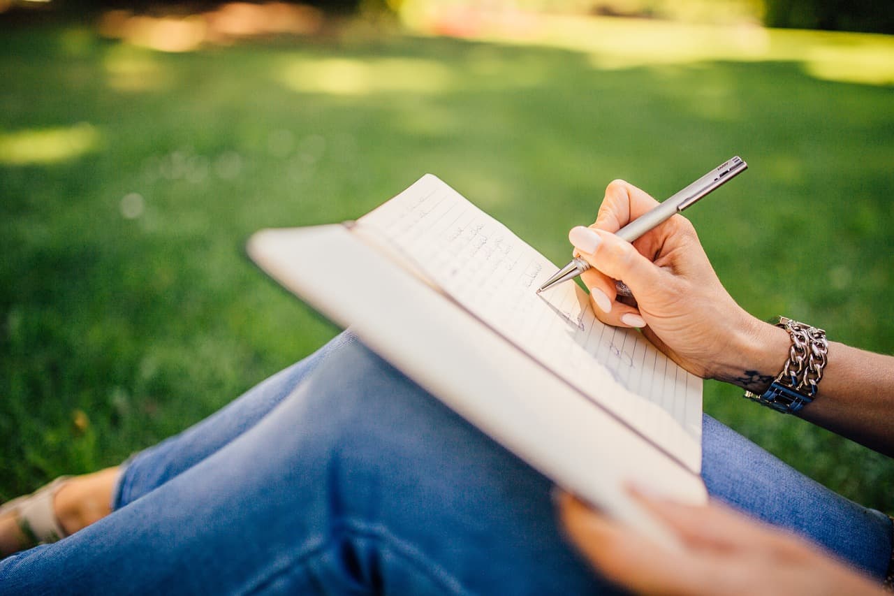 A person sitting on sunlit grass writing notes in a notebook